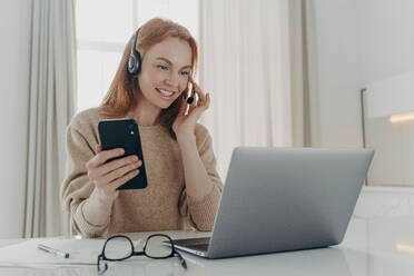 Female employee in headset has video call conference with partners holds modern smartphone watches webinar on laptop computer watches educational lecture poses at desktop against home interior. Female employee in headset has video call conference holds modern smartphone - INGF12517