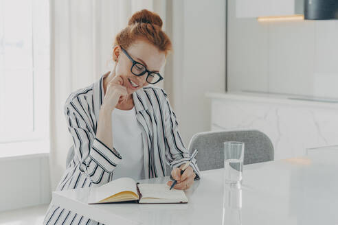 Happy beautiful redhead European woman makes notes in personal planner smiles gently holds pen poses at desktop drinks fresh pure water poses in spacious living room writes memo in her diary.. European woman makes notes in personal planner smiles gently holds pen poses at desktop - INGF12520