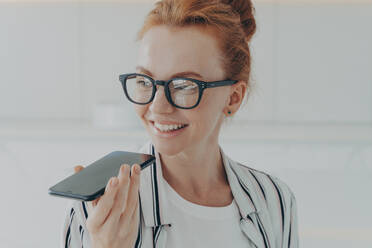 Portrait of happy cheerful red-haired woman in eyewear holding mobile phone, recording audio message and smiling while spending time at home, dressed casually, positive female talking on speakerphone. Portrait of happy cheerful red-haired woman in eyewear holding mobile phone, recording audio message - INGF12521