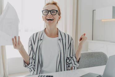 Excited young woman with paper bills in hands feeling euphoric, happy about finally paying down her mortgage debt, exclaiming yes with excitement while sitting at table with laptop. Excited young woman with paper bills in hands finally paying down her mortgage debt at home - INGF12522