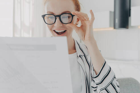 Positive rothaarige junge Frau lächelt breit trägt transparente Brille konzentriert sich auf Papiere prüft Finanzbericht froh, Arbeit in der Zeit zu beenden arbeitet von zu Hause. Menschen Papierkram und Job-Konzept. Positive rothaarige junge Frau lächelt breit trägt transparente Brille konzentriert sich auf Papiere - INGF12523