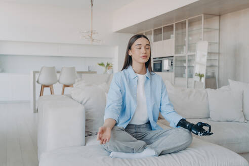 Disabled woman practices morning yoga. Lady in lotus pose on couch, eyes closed. Attractive European girl with myoelectric cyber prosthesis. Modern limb with sensor-based nervous control. - INGF12533