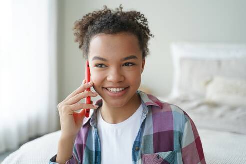 Smiling mixed race teen girl happily answers a phone call, enjoying a pleasant conversation with a friend in the comfort of her bedroom at home. - INGF12538