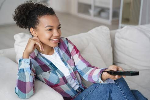 Smiling mixed race teen girl relaxes on the sofa at home, indulging in watching TV series and effortlessly changing TV channels. - INGF12540