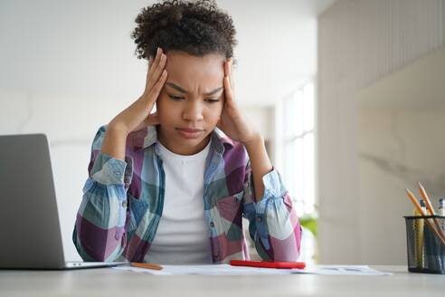 Biracial teen girl school student feels stressed while doing homework on her laptop, facing challenges of difficult e-learning. - INGF12541