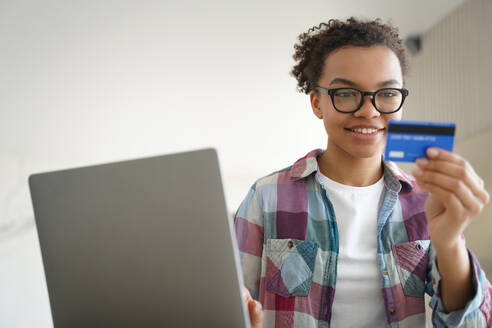 Mixed race teen girl manages her finances, confidently holding a credit card and utilizing online banking services on her laptop for e-commerce purposes at home. - INGF12542