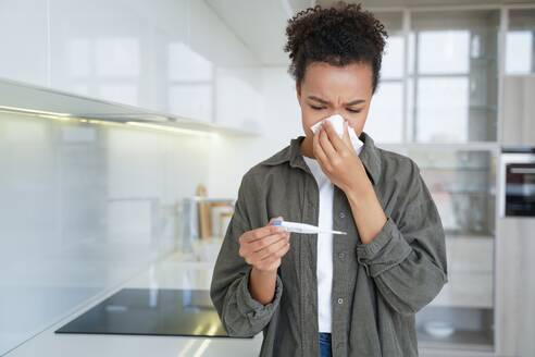 At home, a sick mixed race girl blows her nose and checks her temperature with an electronic thermometer due to flu and fever symptoms. - INGF12546