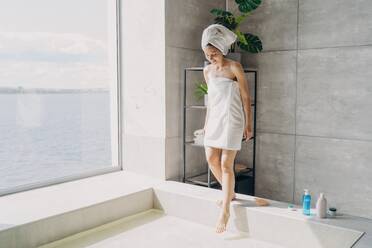 Girl in towels steps into water-filled bath in sunny modern bathroom. Self-care, spa, hygiene. - INGF12548