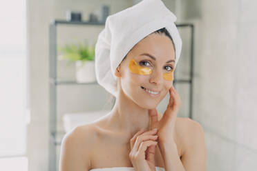 Smiling woman in towel applies golden hydrogel patches under eyes in bathroom. Morning antiaging skincare routine. - INGF12552
