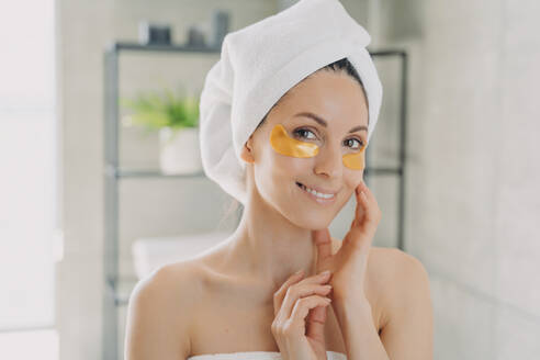 Lächelnde Frau im Handtuch trägt im Badezimmer goldene Hydrogel-Pflaster unter den Augen auf. Morgens Anti-Aging-Pflege. - INGF12552