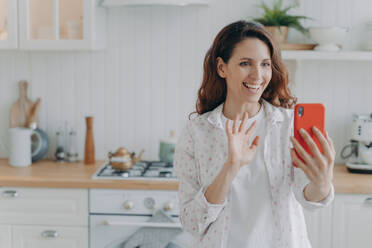 Smiling hispanic female talks on smartphone videocall, waves to camera. Positive woman greets friend, enjoys remote communication in kitchen. - INGF12558