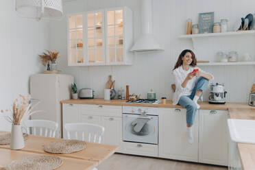 Smiling woman tenant shops online on phone, sitting in modern kitchen. Happy housewife uses smartphone apps, rests after chores. - INGF12559