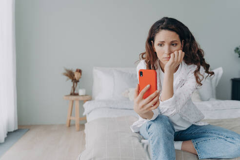 Upset woman reads bad news on smartphone in bedroom. Pensive female shares problems with friend via video call. - INGF12560