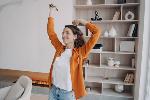 Carefree woman dances with closed eyes, raises hands, listens to music on mobile, enjoying the sound in living room. - INGF12565