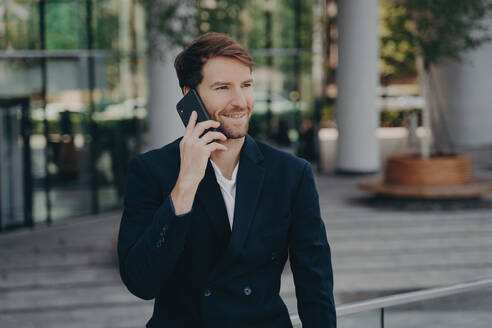 Confident entrepreneur in formal attire makes phone call, enjoying conversation during work break near office building. - INGF12572