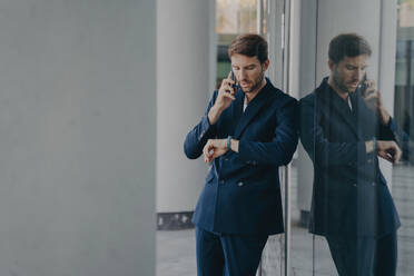 Pensive businessman in suit speaks on phone outside office entrance, glances at watch anxiously, concerned about punctuality for meeting. - INGF12573