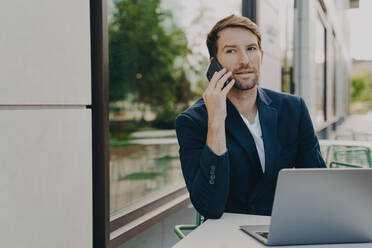 Successful executive consults customer via smartphone, works online with laptop at cafe table, awaiting order. Manager conducts interview. - INGF12574