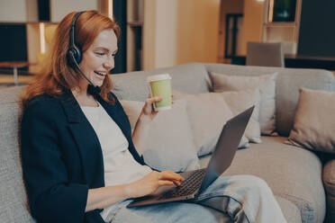 Aufgeregte rothaarige Frau mit drahtlosem Headset und Laptop führt ein Online-Gespräch mit Kollegen, die auf dem Sofa eines Cafés sitzen und die entspannte Atmosphäre bei einem Kaffee genießen. - INGF12575