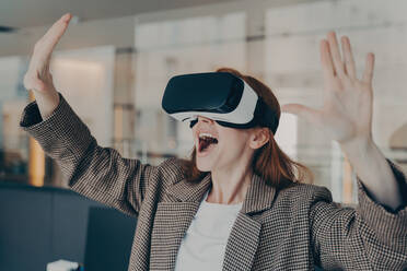 Excited businesswoman in VR glasses, amazed by virtual reality experience in office, expressive gestures and joyful expression. - INGF12577
