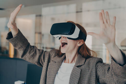 Excited businesswoman in VR glasses, amazed by virtual reality experience in office, expressive gestures and joyful expression. - INGF12577