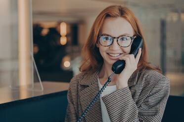 Smiling redhead office worker in plaid jacket and glasses, discussing project completion date on landline phone, office background blurred. - INGF12578