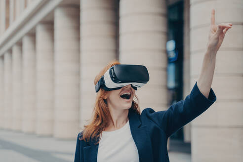 Redhead businesswoman amazed by VR experience outdoors, smiling and attempting to interact with virtual objects, blurred buildings in the background. - INGF12580