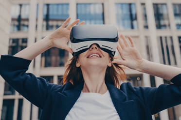 Immersed in virtual reality, futuristic redhead businesswoman wearing 3D goggles stands outside, looking up with awe, holding the portable VR glasses with both hands. - INGF12581