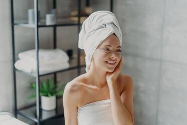 Smiling spa lady with natural makeup poses refreshed after shower, touches healthy skin in bathroom. - INGF12585