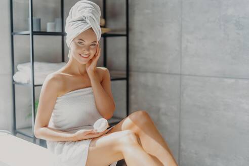 Smiling woman applies facial cream, wrapped in towel, bathroom. - INGF12587