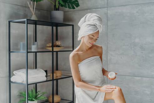 Soft skin after bath. Woman applies cream, wrapped in towel. Beauty and wellness in bathroom. - INGF12588