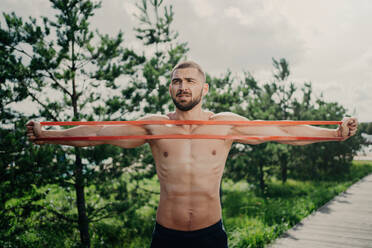 Sporty man with bare chest stretches elastic expander, builds strong muscles, poses outdoors. - INGF12594
