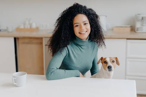 Curly-haired woman in turtleneck sits at kitchen table, enjoys tea, plays with Jack Russell terrier. Joyful moment with beloved pet. - INGF12596