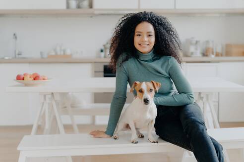 Happy Afro woman sits with dog, poses in kitchen, expresses joy, relaxes with beloved pet. Loving home companionship. - INGF12598