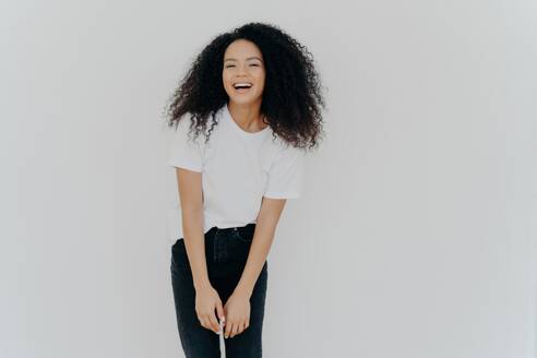 Joyful African American woman in casual wear laughs happily, poses against white background with blank space, feeling energized. - INGF12599