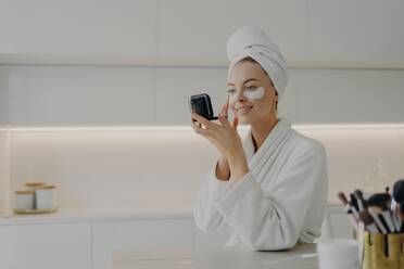 Woman in bathrobe applies under-eye patches, checks mirror in kitchen during morning skincare routine. - INGF12601