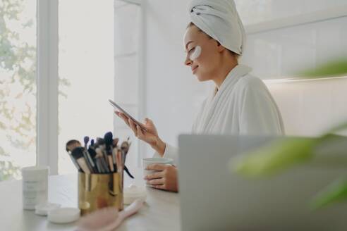 Morning routine: Businesswoman in bathrobe, hair wrapped, under-eye patches, uses smartphone, prepares for work at home, kitchen background. - INGF12602