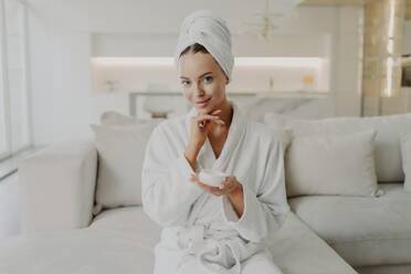 Woman in bathrobe with cream jar, smiles at camera, sits on sofa, does cosmetic procedures, skincare concept. - INGF12606