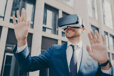 Excited office worker in blue suit outside, using VR glasses to visualize projects, interacting with virtual objects, building in background. - INGF12609