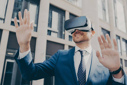 Excited office worker in blue suit outside, using VR glasses to visualize projects, interacting with virtual objects, building in background. - INGF12609