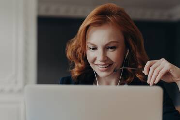Focused freelancer watches tutorial webinar, uses earphones, during online seminar. Happy woman entrepreneur in close-up at laptop. - INGF12612