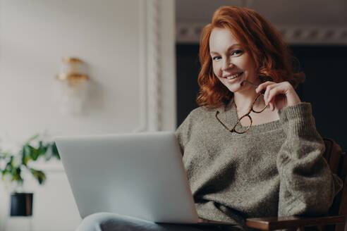 Confident redhead freelancer with spectacles works remotely, plans effective online project, wearing a warm sweater, sitting with laptop. - INGF12621