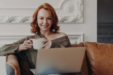 Ginger female on sofa with coffee searches job online, prepares presentation, enjoys cozy atmosphere, high-speed internet. - INGF12622