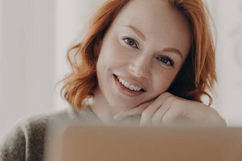 Cheerful ginger woman earns money online, using laptop for freelance work, with a pleasant smile, connected to the internet. - INGF12624