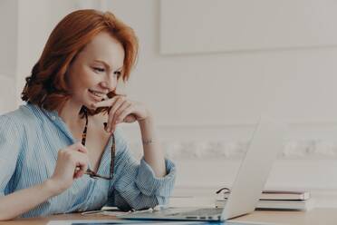 Happy female model at coworking space, with glasses, smiles, uses wireless internet, reads, communicates online, works on web project. - INGF12625