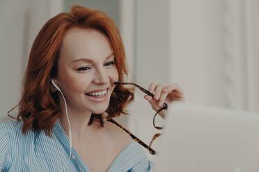 Cheerful redhead woman uses laptop for online conference, discusses work, holds glasses, works freelance from home. Technology concept. - INGF12626