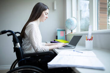 Teenage Girl In Wheelchair Studying At Home On Laptop - INGF12656