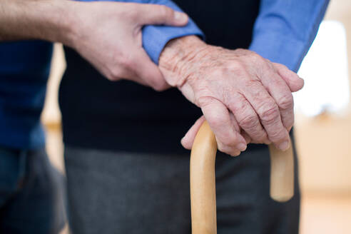 Senior Man’s Hands On Walking Stick With Care Worker In Background - INGF12660