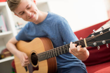 Teenage Boy Sitting On Sofa At Home Playing Guitar - INGF12665