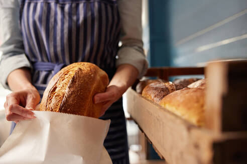 Sales Assistant In Bakery Putting Freshly Baked Organic Sourdough Bread Loaf Into Sustainable Paper Bag - INGF12672