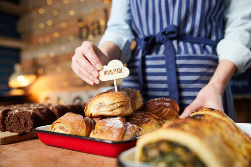 Sales Assistant In Bakery Putting Homemade Label Into Stack Of Freshly Baked Baked Cinnamon Buns - INGF12674